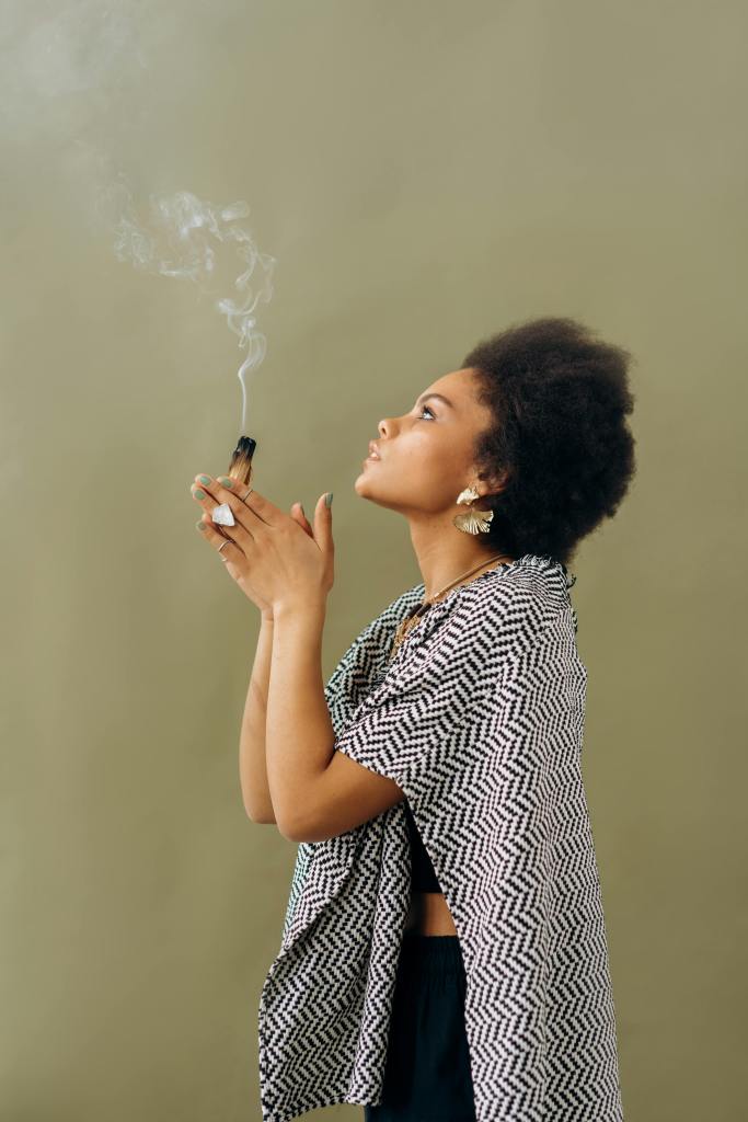 Woman with her hands together in front of her face holding a partial burnt piece of wood with a thin stream of smoke going up.