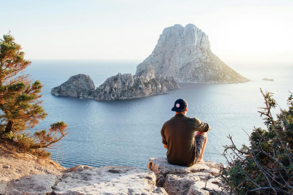 Man siting on some rocks looking out at an open body of water with another large rock structure in the middle.